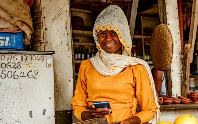 Kenyan shopkeeper using her phone at her shop counter, representing small business compliance with eTIMS electronic tax invoicing