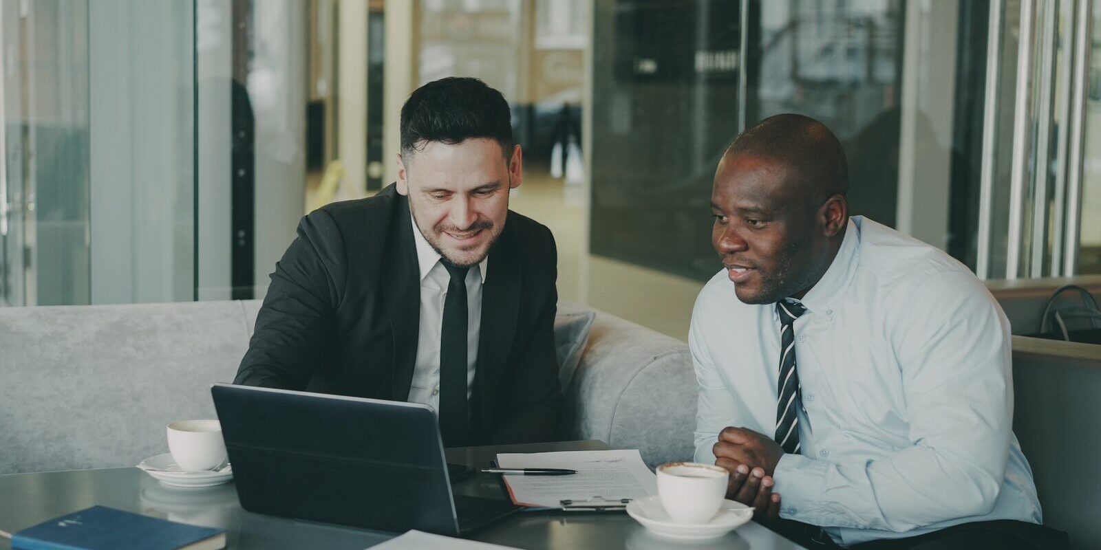 Two African businessmen reviewing financial documents with a laptop and coffee, discussing profit repatriation structuring