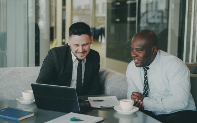 Two African businessmen reviewing financial documents with a laptop and coffee, discussing profit repatriation structuring