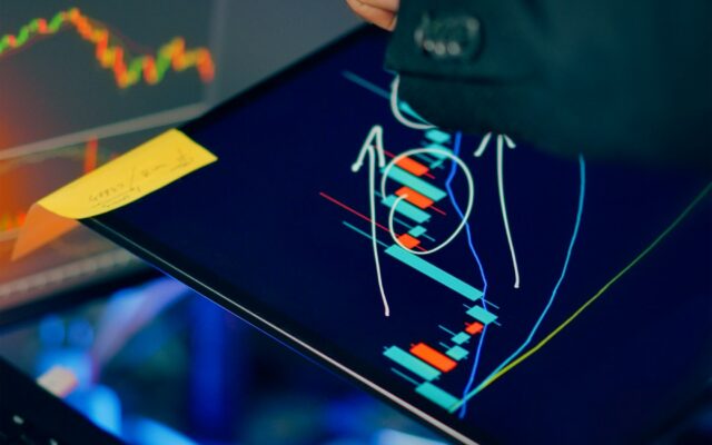 A hand pointing at financial charts on a tablet and screens