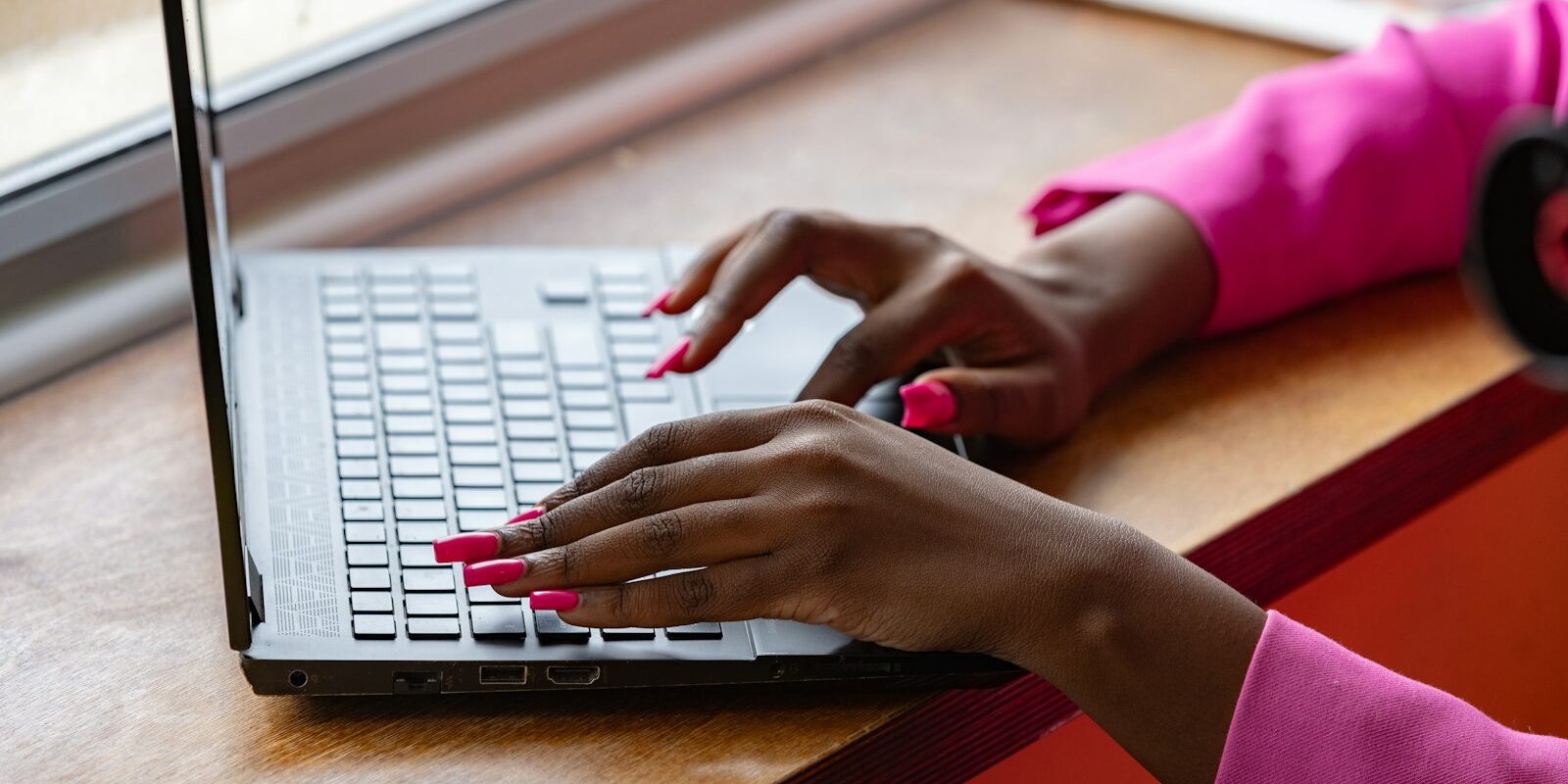 Hands typing on a laptop keyboard at a wooden desk by a window